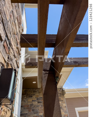 An outdoor residential pergola with exposed dark wood beams and a stone exterior wall. The image is captured looking upward, showing the clear blue sky between the architectural elements. 128421298