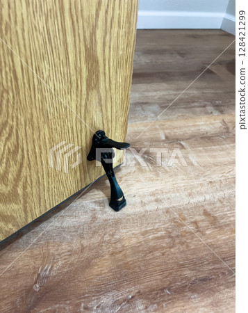 Close-up of a black kickdown doorstop installed under a wooden door in a room with medium-tone laminate flooring. 128421299