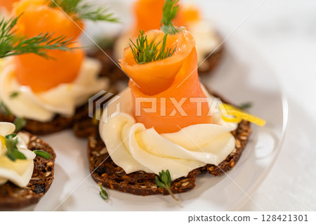 A gourmet smoked salmon canape elegantly arranged on a crispy cracker, topped with a swirl of smooth cream cheese, fresh thyme, and a drizzle of olive oil. The vibrant orange salmon contrasts 128421301