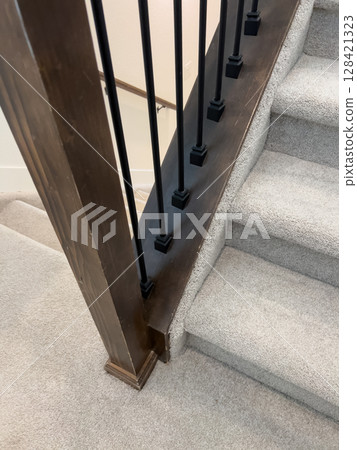 Close-up of the base of a staircase with a wood post, black spindles, and beige carpeted steps. The image highlights the structural transition between materials in a modern home. 128421323