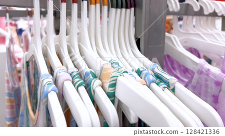 Colorful children's summer dresses with striped and tropical prints hang on white plastic hangers. The rack is organized and placed in a well-lit retail space. Colorful children's summer dresses with striped and tropical prints hang on white plastic hangers. The rack is organized and placed in a well-lit retail space. 128421336