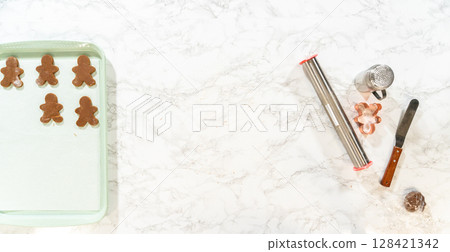 Overhead view of a baking tray with neatly placed gingerbread cookies, dough remnants, and baking tools on a marble surface. Overhead view of a baking tray with neatly placed gingerbread cookies, dough remnants, and baking tools on a marble surface. 128421342