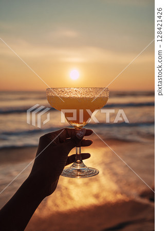 Woman's hand holding a cocktail on the beach at sunset 128421426
