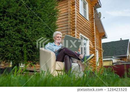 A senior woman is engaged in her work outdoors, using a laptop in a cozy garden environment 128423320