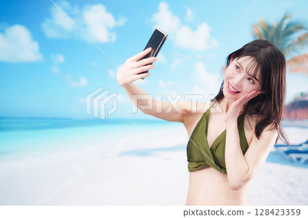 一位身穿泳衣的年輕女子在陽光明媚的海灘上用智慧型手機自拍 128423359