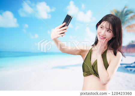 一位身穿泳衣的年輕女子在陽光明媚的海灘上用智慧型手機自拍 128423360