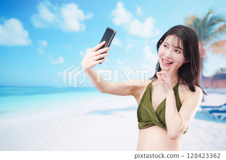 一位身穿泳衣的年輕女子在陽光明媚的海灘上用智慧型手機自拍 128423362
