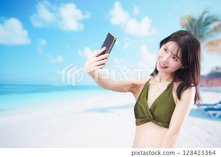 A young woman in a swimsuit taking a selfie with her smartphone on a sunny beach 128423364