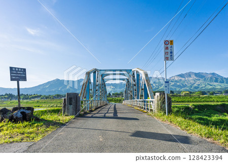 The "Kaizegawa Bridge" in Minamiaso Village and rural scenery with the blue sky and the five peaks of Mt. Aso in the background. A bridge in the river in Minamiaso Village. The "Kaizegawa Bridge" in Minamiaso Village and rural scenery with the blue sky and the five peaks of Mt. Aso in the background. A bridge in the river in Minamiaso Village. 128423394
