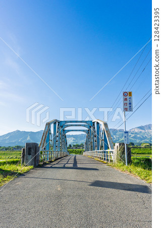 The "Kaizegawa Bridge" in Minamiaso Village and rural scenery with the blue sky and the five peaks of Mt. Aso in the background. A bridge in the river in Minamiaso Village. The "Kaizegawa Bridge" in Minamiaso Village and rural scenery with the blue sky and the five peaks of Mt. Aso in the background. A bridge in the river in Minamiaso Village. 128423395