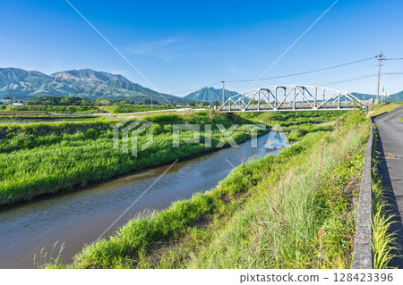 The "Kaizegawa Bridge" in Minamiaso Village and rural scenery with the blue sky and the five peaks of Mt. Aso in the background. A bridge in the river in Minamiaso Village. The "Kaizegawa Bridge" in Minamiaso Village and rural scenery with the blue sky and the five peaks of Mt. Aso in the background. A bridge in the river in Minamiaso Village. 128423396