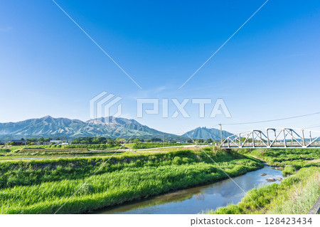 The "Kaizegawa Bridge" in Minamiaso Village and rural scenery with the blue sky and the five peaks of Mt. Aso in the background. A bridge in the river in Minamiaso Village. The "Kaizegawa Bridge" in Minamiaso Village and rural scenery with the blue sky and the five peaks of Mt. Aso in the background. A bridge in the river in Minamiaso Village. 128423434