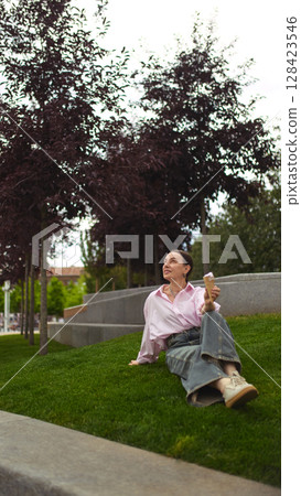 Calm sweetness. Elderly woman in pink shirt sitting on grass with ice cream cone in peaceful summer park Calm sweetness. Elderly woman in pink shirt sitting on grass with ice cream cone in peaceful summer park 128423546