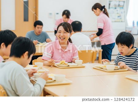 一位面帶微笑的女護理員在殘疾人設施內為殘疾人士提供餐食。提供食物、午餐時間、學校午餐、餐點、樂趣 一位面帶微笑的女護理員在殘疾人設施內為殘疾人士提供餐食。提供食物、午餐時間、學校午餐、餐點、樂趣 128424172