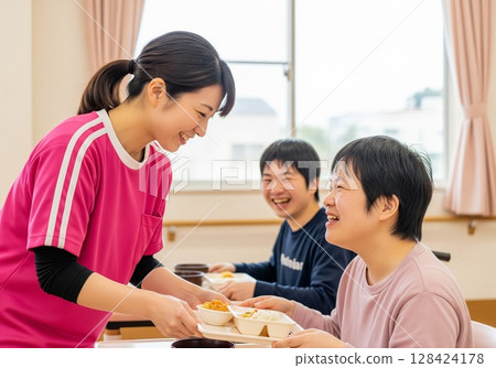 一位面帶微笑的女護理員在殘疾人設施內為殘疾人士提供餐食。提供食物、午餐時間、學校午餐、餐點、樂趣 128424178