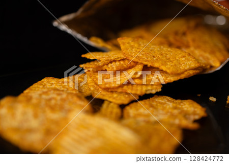 Golden-orange ridged potato chips spill from an open foil bag, highlighting their crunchy texture as an indulgent snack, perfect for casual eating Golden-orange ridged potato chips spill from an open foil bag, highlighting their crunchy texture as an indulgent snack, perfect for casual eating 128424772