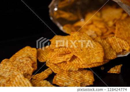Golden crunchy ridged potato chips spilling from a bag onto a dark background, symbolizing delicious indulgent snack or unhealthy food choice Golden crunchy ridged potato chips spilling from a bag onto a dark background, symbolizing delicious indulgent snack or unhealthy food choice 128424774