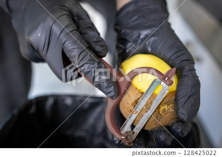 Person's hands wearing black safety gloves expertly peeling a fresh brown potato with a kitchen peeler, emphasizing food preparation and culinary hygiene 128425022