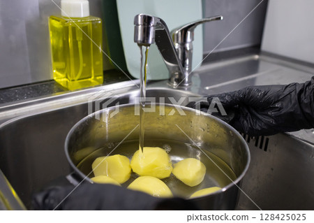 Person in black gloves washes fresh peeled potatoes in a metal pot under running water at a commercial kitchen sink, promoting culinary hygiene and food preparation Person in black gloves washes fresh peeled potatoes in a metal pot under running water at a commercial kitchen sink, promoting culinary hygiene and food preparation 128425025