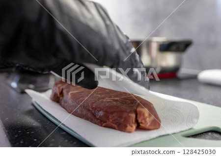 Professional chefs gloved hand gently patting dry raw beef steak with paper towel on cutting board for hygienic home kitchen food preparation 128425030