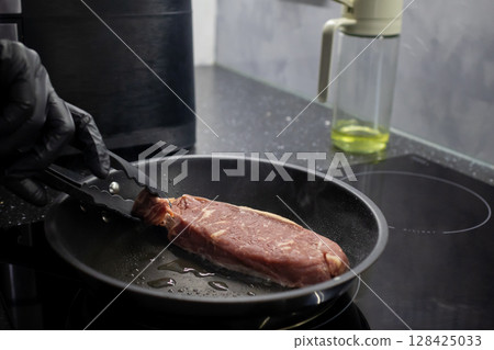 Professional chef in black gloves cooking raw beef steak in a hot frying pan on a modern induction cooktop for gourmet meal preparation Professional chef in black gloves cooking raw beef steak in a hot frying pan on a modern induction cooktop for gourmet meal preparation 128425033