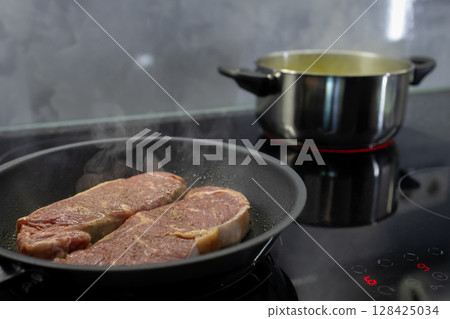 Two raw meat steaks sizzling in a hot frying pan on a modern induction hob, showcasing delicious home cooking, meal preparation, and culinary activity Two raw meat steaks sizzling in a hot frying pan on a modern induction hob, showcasing delicious home cooking, meal preparation, and culinary activity 128425034
