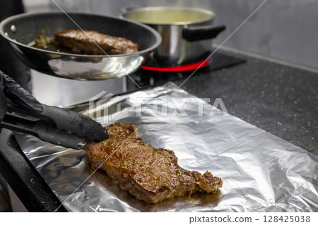 Skilled gloved hand transferring a perfectly seared steak from a hot frying pan to aluminum foil using tongs, symbolizing gourmet home cooking Skilled gloved hand transferring a perfectly seared steak from a hot frying pan to aluminum foil using tongs, symbolizing gourmet home cooking 128425038