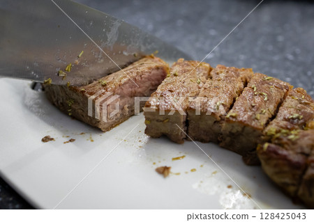 A sharp kitchen knife expertly slicing a juicy cooked beef tenderloin roast on a white cutting board, highlighting delicious culinary preparation for a healthy meal A sharp kitchen knife expertly slicing a juicy cooked beef tenderloin roast on a white cutting board, highlighting delicious culinary preparation for a healthy meal 128425043