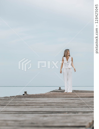 Asian woman in white outfit stands on wooden pier, calm lake, peaceful overcast, viewed from behind with contemplative mood, mindfulness, travel retreats, wellness, vertical, copy space Asian woman in white outfit stands on wooden pier, calm lake, peaceful overcast, viewed from behind with contemplative mood, mindfulness, travel retreats, wellness, vertical, copy space 128425045