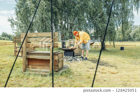 man in red cap, in yellow clothes near outdoor fire pit surrounded by stones with rustic benches seen through tent ropes, camping equipment, travel agency, outdoor adventure man in red cap, in yellow clothes near outdoor fire pit surrounded by stones with rustic benches seen through tent ropes, camping equipment, travel agency, outdoor adventure 128425051