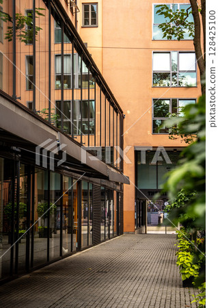 Modern Street Scene In Munich Center: Contemporary Architecture With Glass Facades And Greenery In A Quiet Urban Courtyard 128425100
