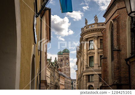 Iconic Cathedral Of Our Dear Lady In Munich: Majestic Twin Domes Rising Above Historic Buildings Under A Bright Blue Sky With Scenic Clouds Near Marienplatz 128425101