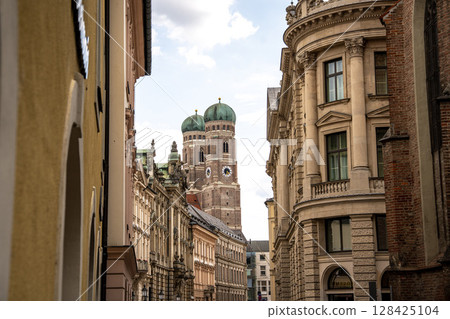 Iconic Cathedral Of Our Dear Lady In Munich: Majestic Twin Domes Rising Above Historic Buildings Under A Bright Blue Sky With Scenic Clouds Near Marienplatz Iconic Cathedral Of Our Dear Lady In Munich: Majestic Twin Domes Rising Above Historic Buildings Under A Bright Blue Sky With Scenic Clouds Near Marienplatz 128425104