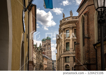 Iconic Cathedral Of Our Dear Lady In Munich: Majestic Twin Domes Rising Above Historic Buildings Under A Bright Blue Sky With Scenic Clouds Near Marienplatz 128425106