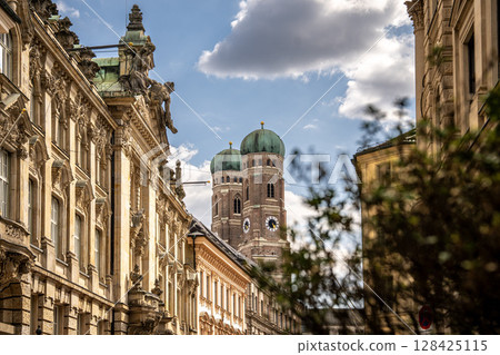 Iconic Cathedral Of Our Dear Lady In Munich: Majestic Twin Domes Rising Above Historic Buildings Under A Bright Blue Sky With Scenic Clouds Near Marienplatz 128425115