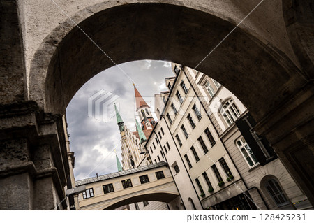 Historic Toy Museum In Munich Near Marienplatz: Old Tower With Red Roof And Green Spire Framed By Stone Arch Against Dramatic Cloudy Sky 128425231