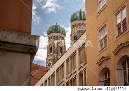 Iconic Cathedral Of Our Dear Lady In Munich: Majestic Twin Domes Rising Above Historic Buildings Under A Bright Blue Sky With Scenic Clouds Near Marienplatz Iconic Cathedral Of Our Dear Lady In Munich: Majestic Twin Domes Rising Above Historic Buildings Under A Bright Blue Sky With Scenic Clouds Near Marienplatz 128425234