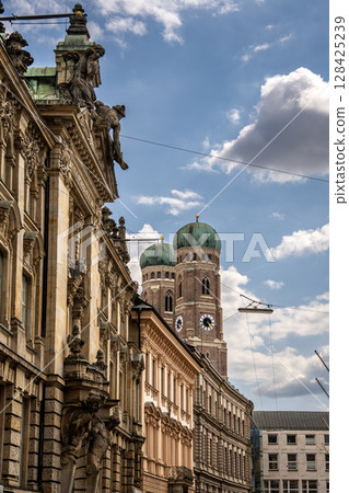 Iconic Cathedral Of Our Dear Lady In Munich: Majestic Twin Domes Rising Above Historic Buildings Under A Bright Blue Sky With Scenic Clouds Near Marienplatz 128425239