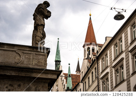 Historic Toy Museum In Munich Near Marienplatz: Old Tower With Red Roof And Green Spire Framed By Stone Arch Against Dramatic Cloudy Sky Historic Toy Museum In Munich Near Marienplatz: Old Tower With Red Roof And Green Spire Framed By Stone Arch Against Dramatic Cloudy Sky 128425288
