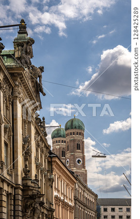 Iconic Cathedral Of Our Dear Lady In Munich: Majestic Twin Domes Rising Above Historic Buildings Under A Bright Blue Sky With Scenic Clouds Near Marienplatz Iconic Cathedral Of Our Dear Lady In Munich: Majestic Twin Domes Rising Above Historic Buildings Under A Bright Blue Sky With Scenic Clouds Near Marienplatz 128425289