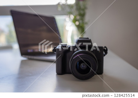 Modern digital camera with a prominent lens on a clean white desk, showcasing professional photography equipment and creative digital content creation tools with a laptop 128425514