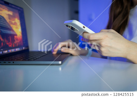 Woman's hands operate laptop and smartphone simultaneously, depicting digital multitasking for modern remote work and smart home technology management Woman's hands operate laptop and smartphone simultaneously, depicting digital multitasking for modern remote work and smart home technology management 128425515