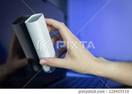 Human hand diligently cleans a reflective laptop screen with a modern white electronic device cleaning tool, highlighting essential digital hygiene and tech care for optimal performance 128425519