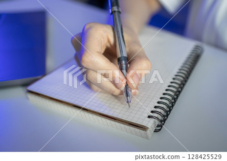 A person's hand holds a sleek pen, diligently writing in a checkered spiral notebook, symbolizing focus, learning, and productivity for education or professional work 128425529