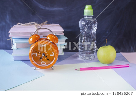 On the table is a school lunch, an alarm clock and stationery. A sandwich, an apple and a bottle of water. School lunch and textbooks. On the table is a school lunch, an alarm clock and stationery. A sandwich, an apple and a bottle of water. School lunch and textbooks. 128426207