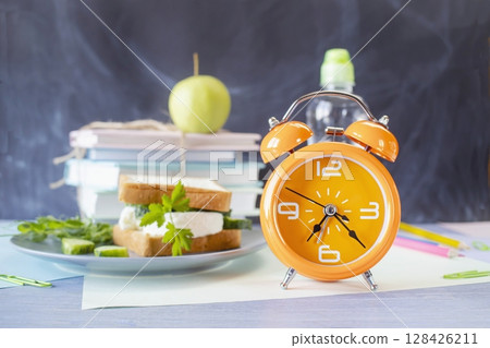 On the table is a school lunch, an alarm clock and stationery. A sandwich, an apple and a bottle of water. School lunch and textbooks. 128426211