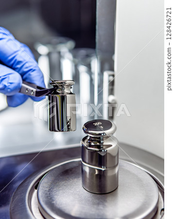 A operator's hand is holding steel calibration weight to place on the analytical balance. A operator's hand is holding steel calibration weight to place on the analytical balance. 128426721