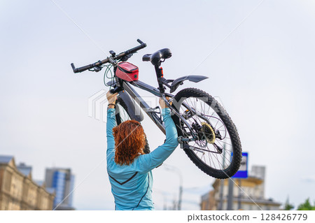 Woman holding bicycle triumphantly above head against clear sky after urban ride, celebrating success and power. Concept of cycling, freedom, cycling, personal sport achievement outdoors Woman holding bicycle triumphantly above head against clear sky after urban ride, celebrating success and power. Concept of cycling, freedom, cycling, personal sport achievement outdoors 128426779