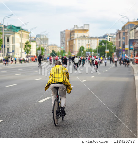 Cyclist in yellow checkered jacket rides down a wide city street among a group of bikers during an urban cycling event. Concept of city biking, sustainable transport, active lifestyle 128426788