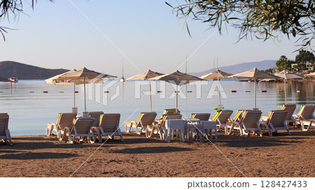 Beach chairs and umbrellas line a sandy shore, facing a calm sea with boats and distant hills under a clear sky. High quality photo Beach chairs and umbrellas line a sandy shore, facing a calm sea with boats and distant hills under a clear sky. High quality photo 128427433